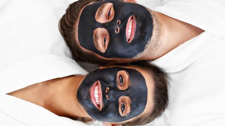 Two spa clients lying side by side wearing black charcoal facial masks on white towels during a couples' treatment — illustrating how ScanSkinAI recommends the right in-spa service for each client's scan result.